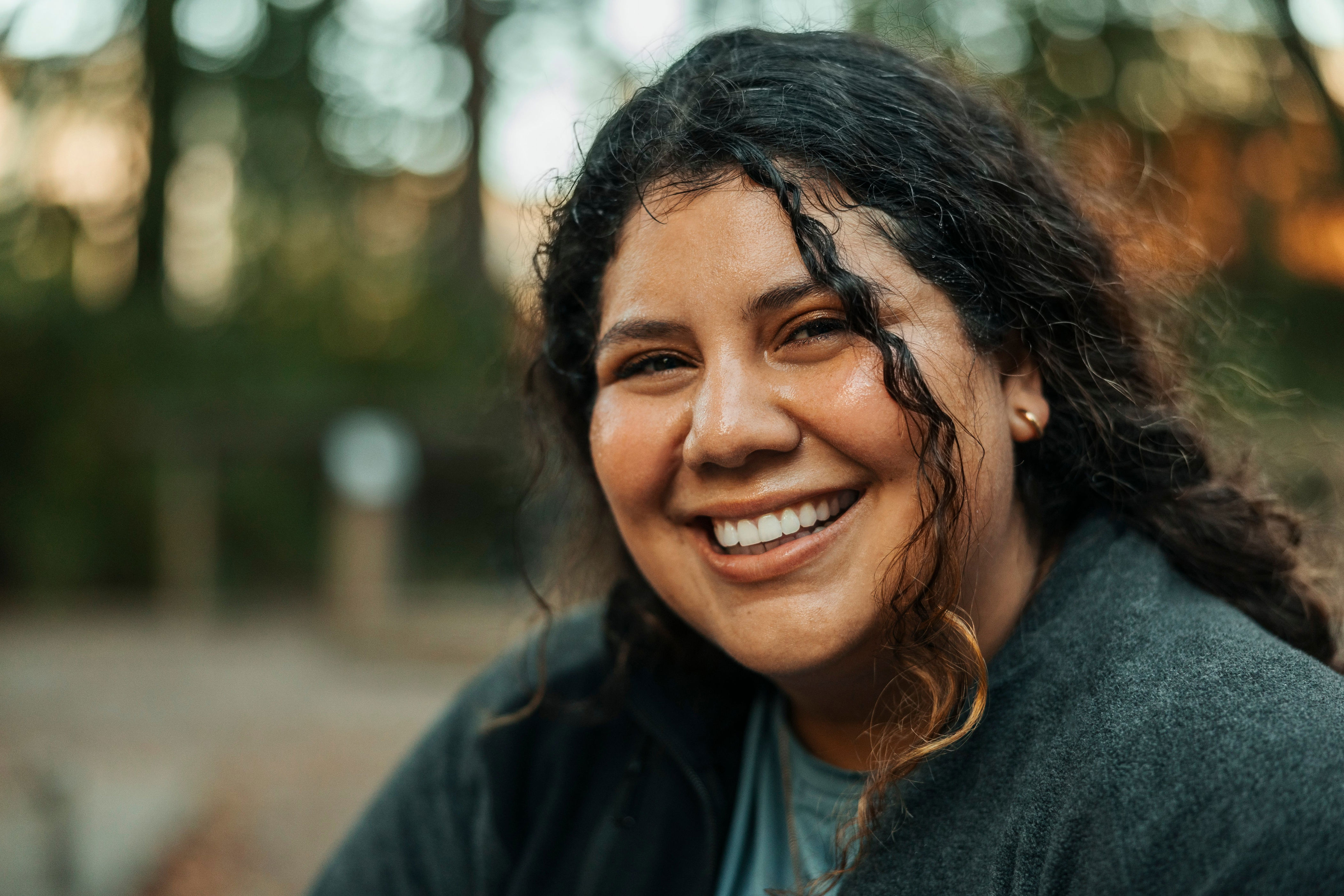 Smiling person outdoors, wearing a casual dark jacket. Curly hair frames their face as they look directly at the camera