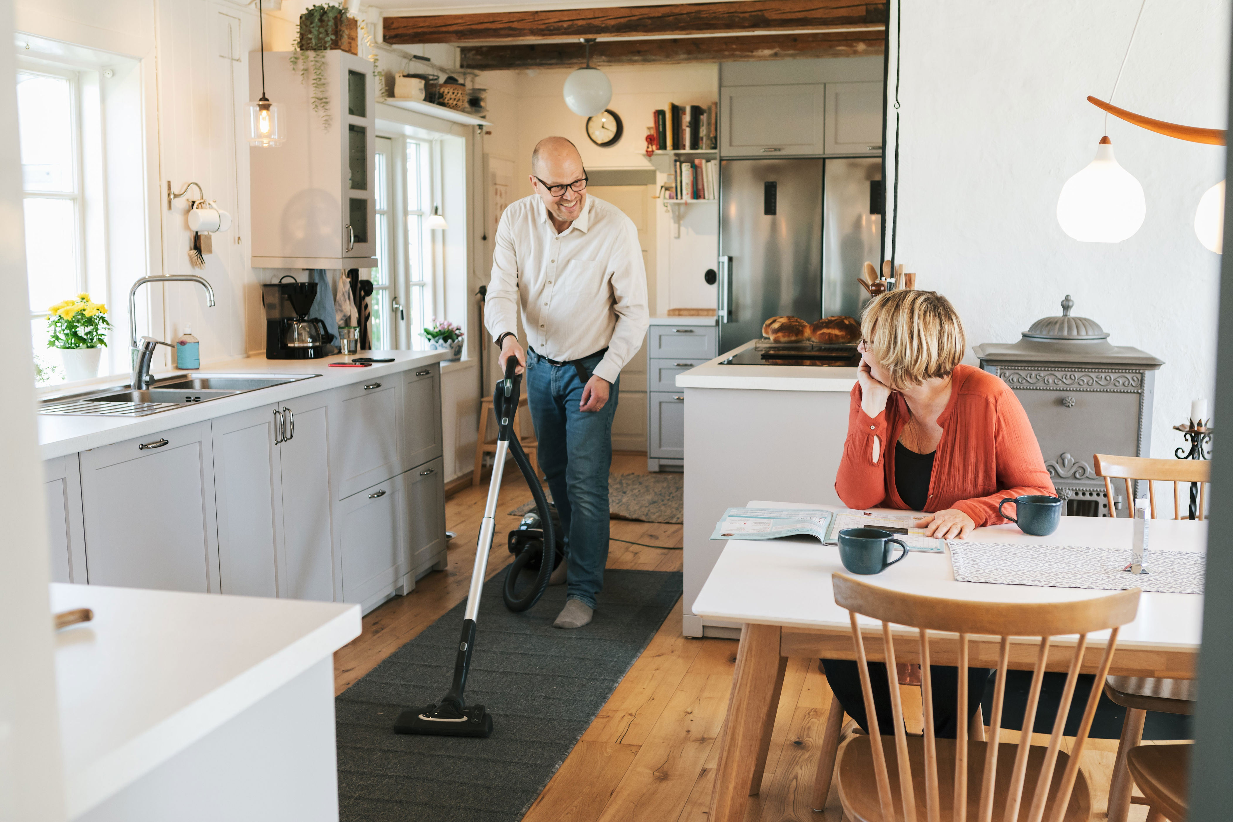 Man vacuuming in a kitchen while a woman reads at a table nearby. The setting is a bright, tidy home
