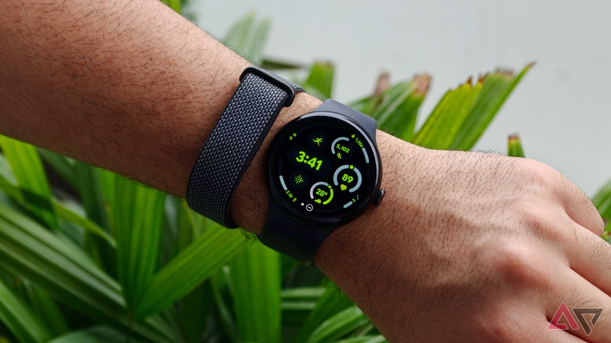 Close-up of a person's wrist wearing a Google Pixel Watch with a black silicone band alongside the Fitbit Air against a green leafy background