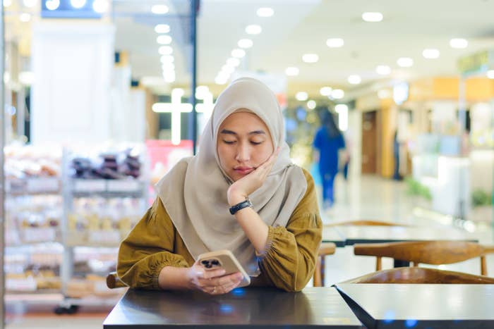 A person in a hijab intently looks at their smartphone while seated at a table in a well-lit shopping area