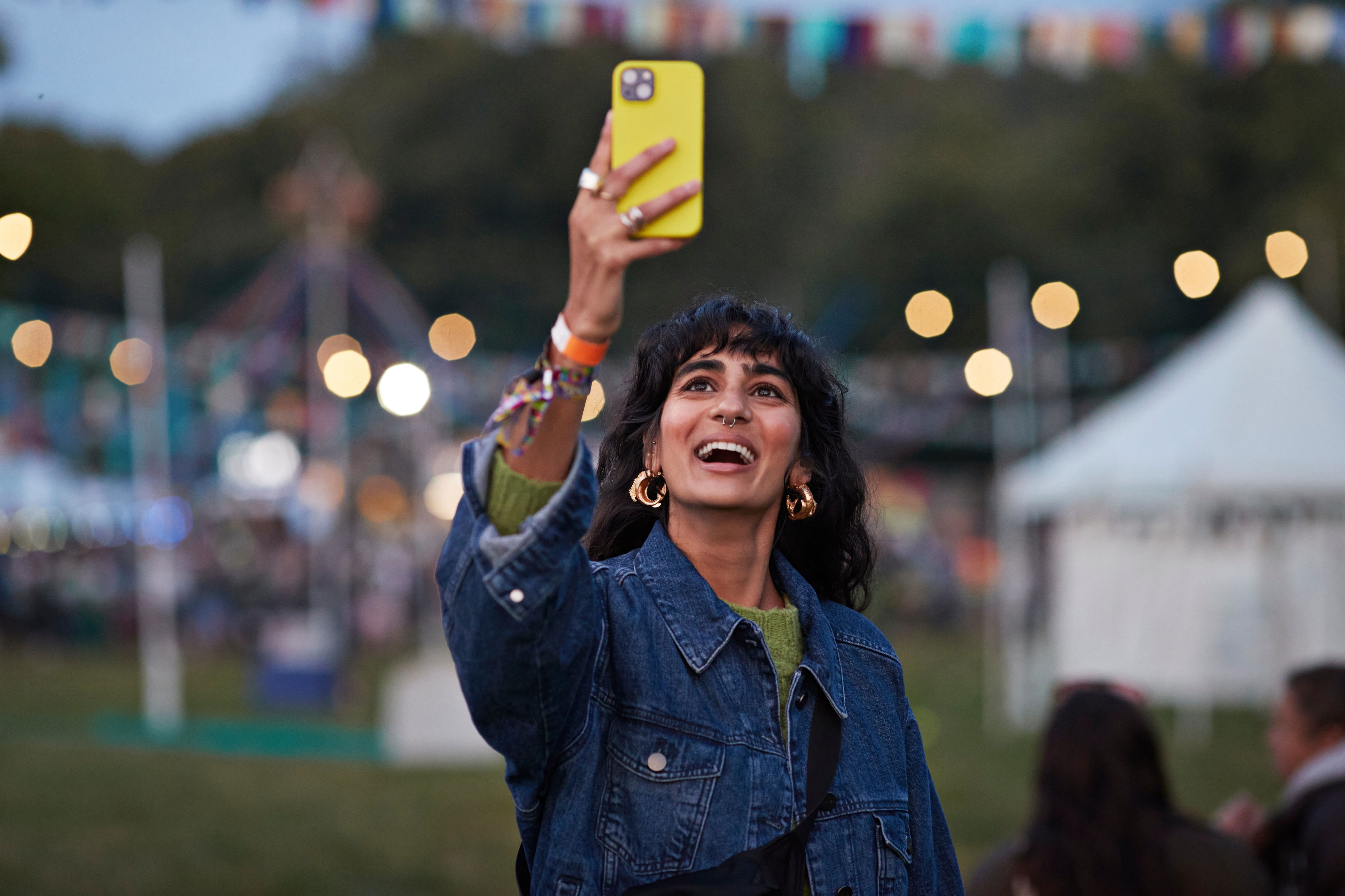 A person joyfully takes a selfie at an outdoor festival, wearing a denim jacket and gold hoop earrings. String lights and tents are visible in the background