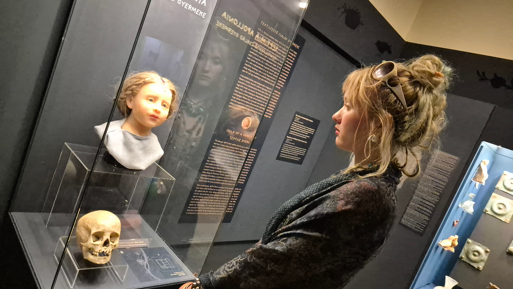A woman stands in front of a glass display case showing a young face and a skull in front of it