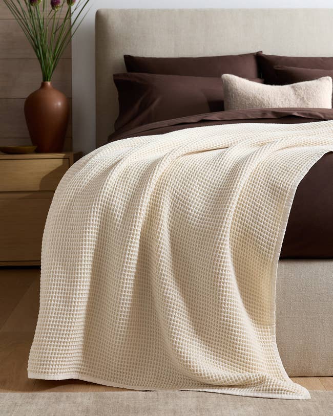 Cream waffle-knit throw draped over a neatly made bed with brown bedding and decorative pillows, creating a cozy and inviting look