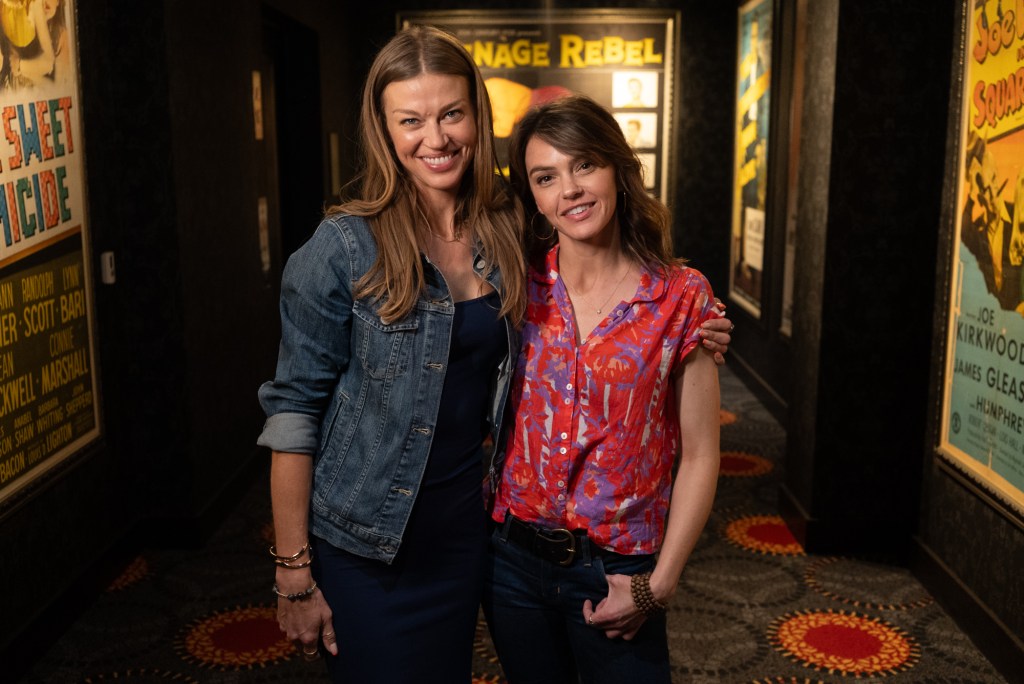 Adrianne Palicki and Aimee Teegarden smiling in a hallway with movie posters.