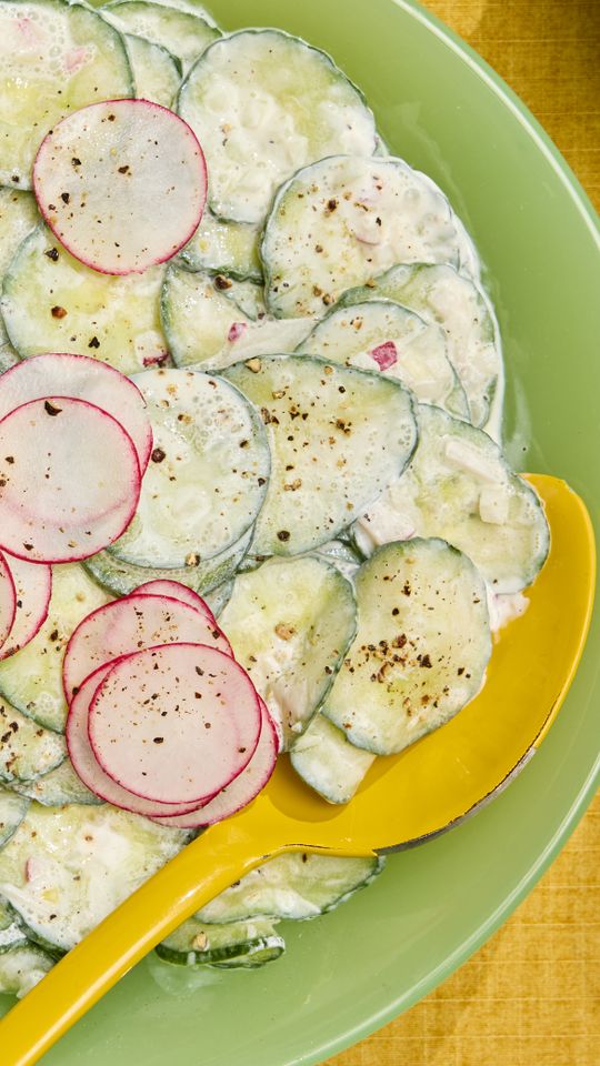 cucumbers and radishes in a seasoned, white sour cream sauce with a spoon on a green plate and yellow background