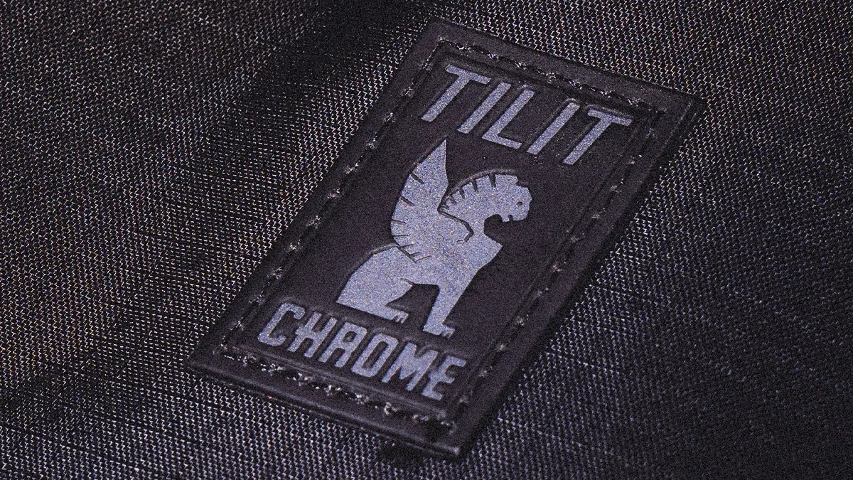 Black fabric with a stitched rectangular patch featuring a winged lion logo and the text