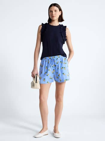 Person in a sleeveless ruffled top, pineapple print shorts, and flats, holding a woven bag, stands against a plain background