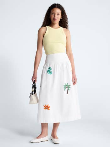 Person wearing a sleeveless top and a midi skirt with botanical embroidery, holding a small bag, posing in a studio setting