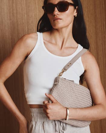 Model in a white tank top and sunglasses holding a woven handbag, styled for a casual yet chic look