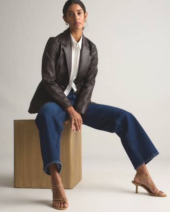 A person sits on a wooden block, wearing a blazer over a white shirt, wide-leg jeans, and heels