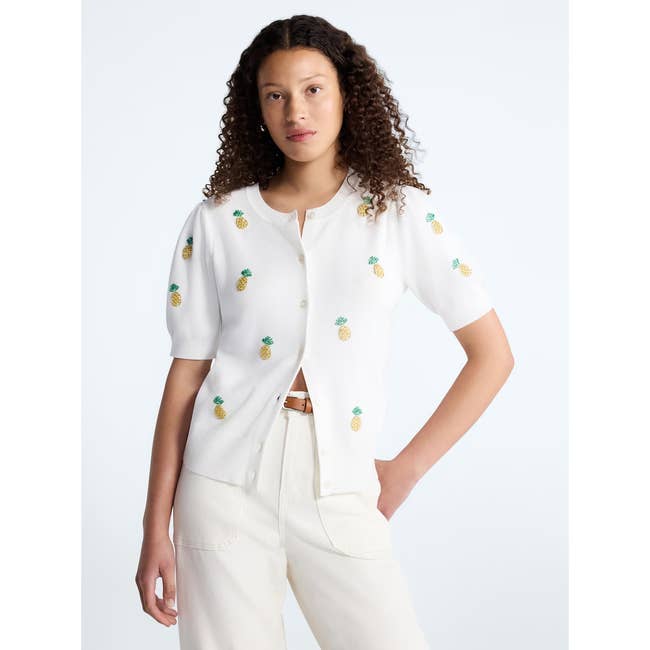 Person wearing a white cardigan with small pineapple patterns, buttoned partially, paired with matching white pants