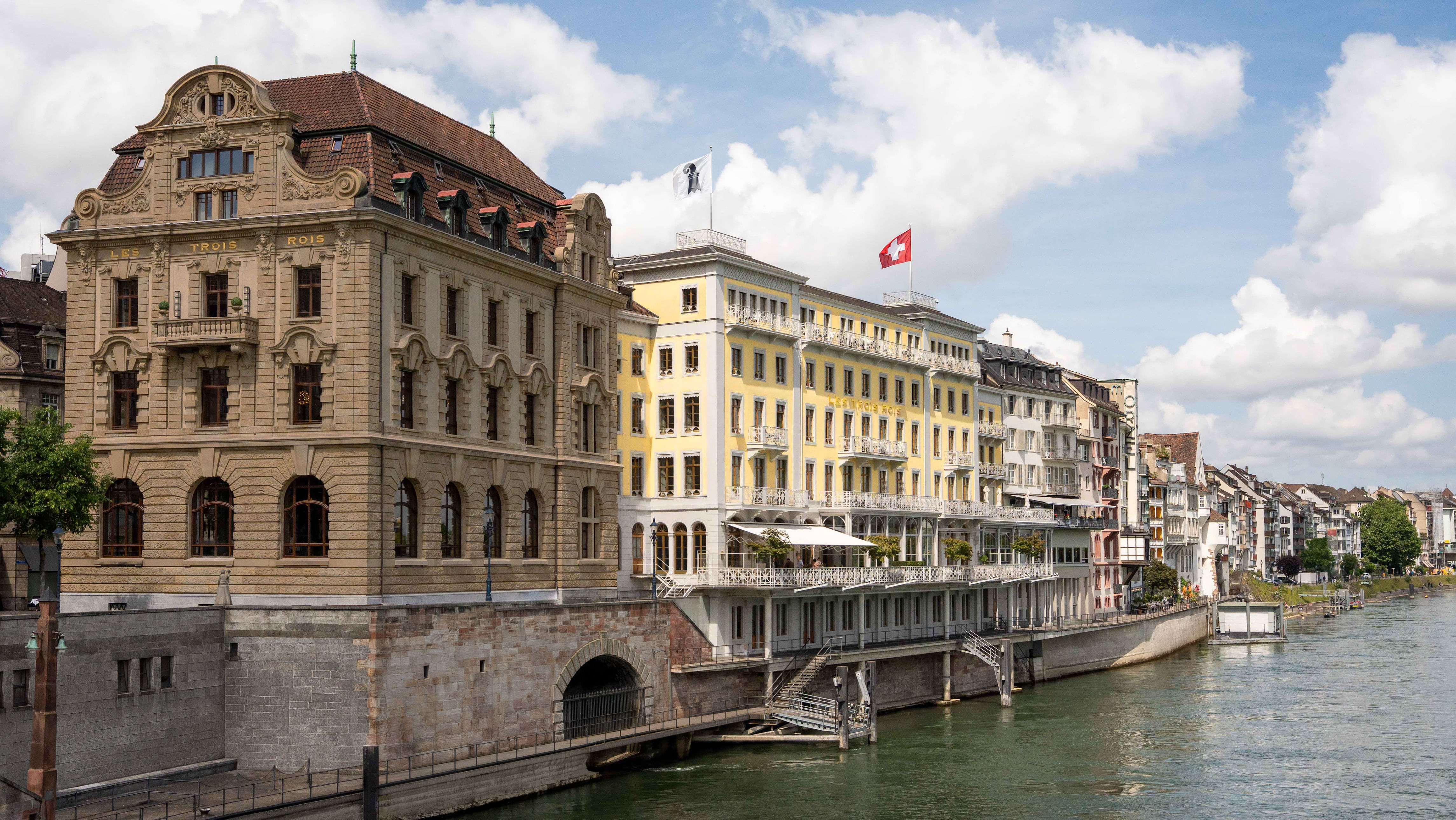 The exterior of Les Trois Rois, a brown building next to a yellow building with white balconies, along the Rhine, with a red and white Switzerland flag on top.
