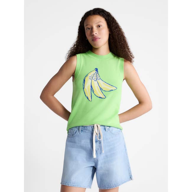 Person in sleeveless top with banana print and denim shorts stands confidently