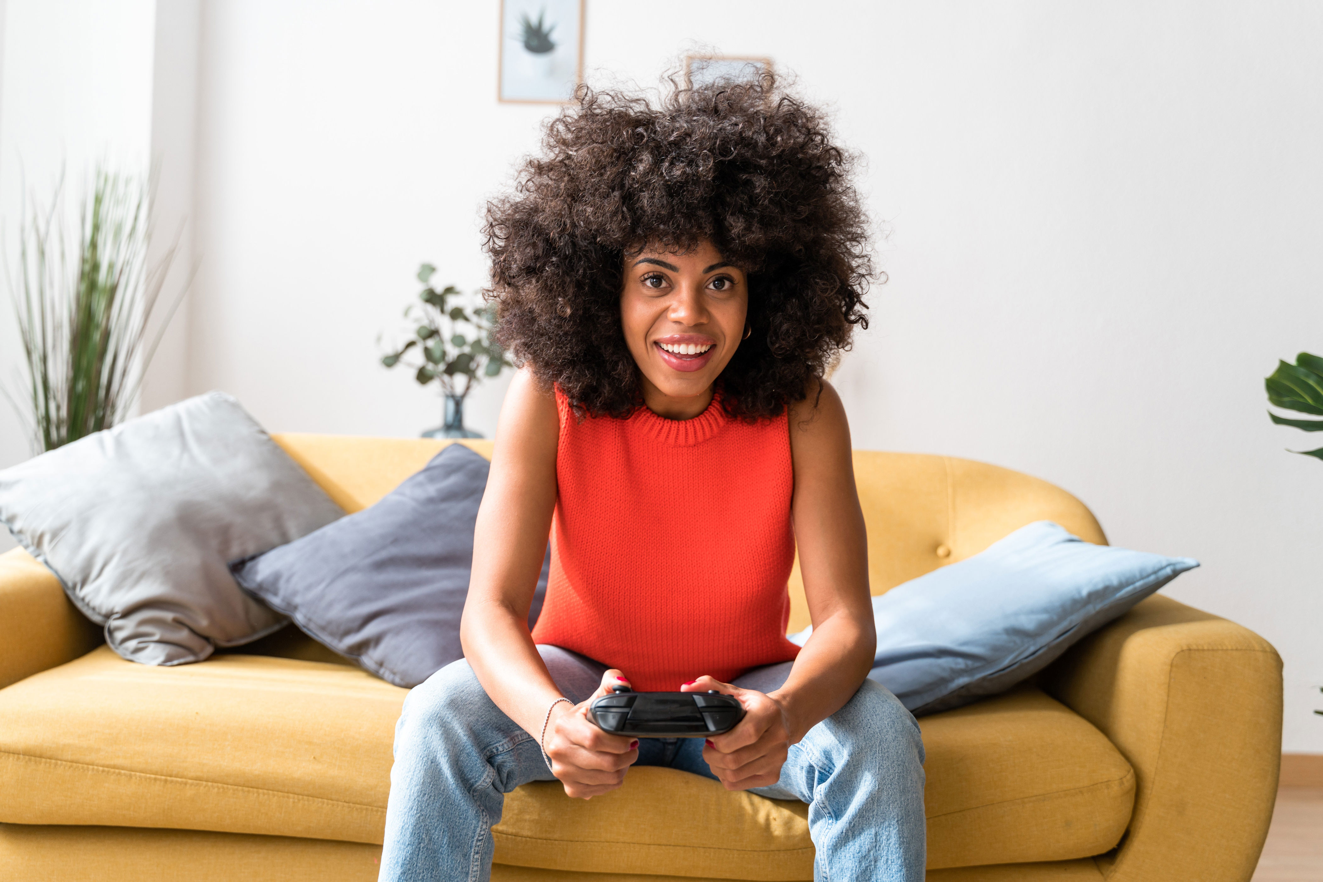 Person with curly hair smiling while holding a game controller, sitting on a couch with cushions in a living room setting