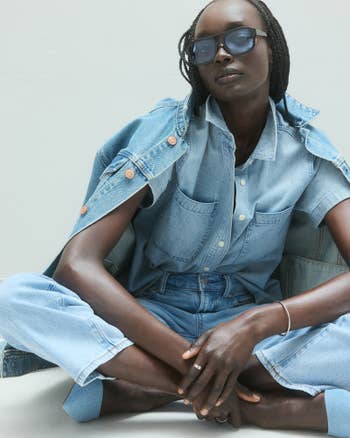 Person wearing a stylish denim outfit with sunglasses, sitting cross-legged