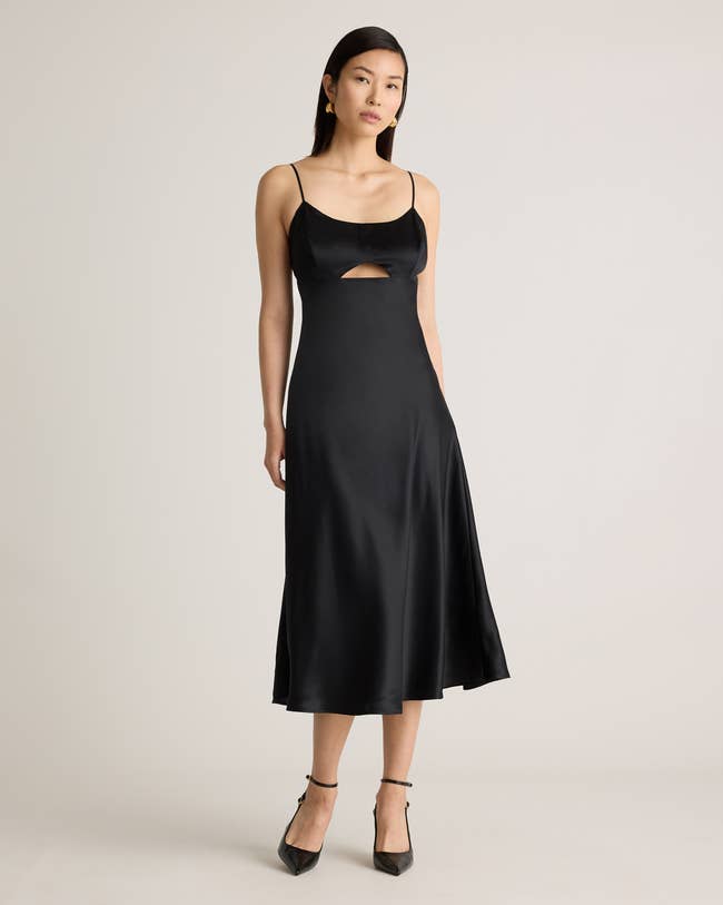 A person in a sleek, sleeveless black satin dress with a front cutout, standing in a neutral pose