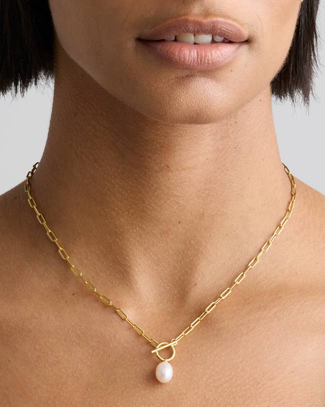 A close-up of a paperclip chain necklace with a toggle clasp and single pearl pendant worn around a woman's neck