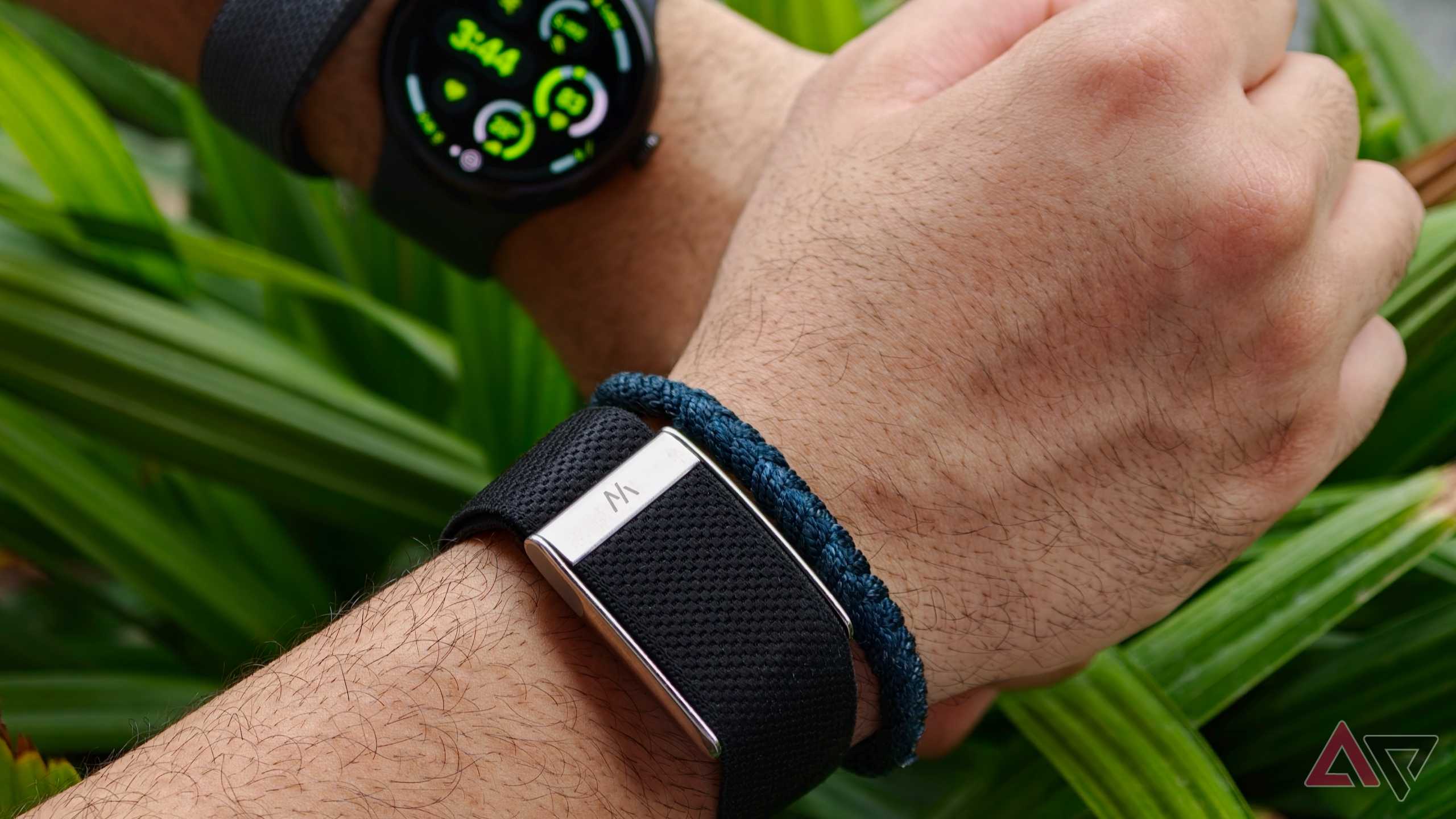Close-up focus on a Whoop fitness tracker band on a wrist with its metallic clasp visible, while a Google Pixel Watch is blurred in the background