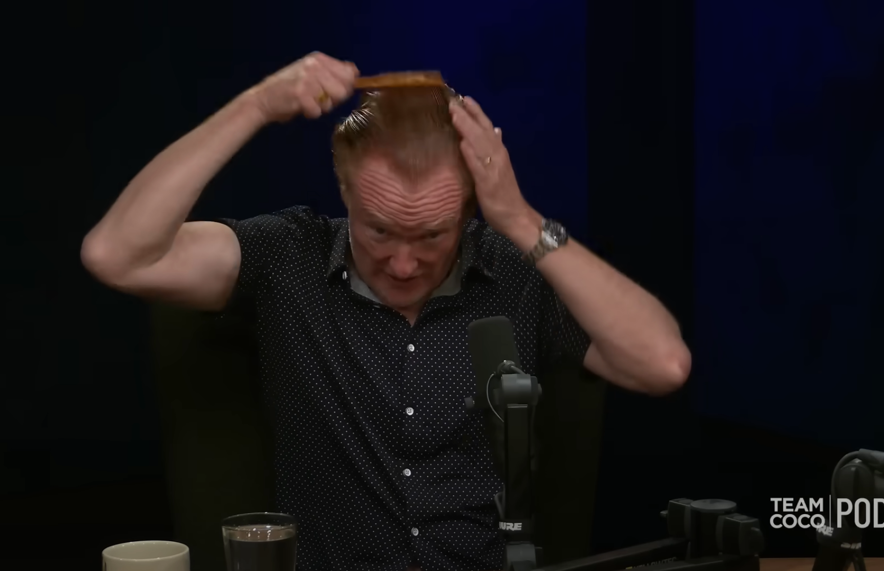 Person brushing hair during a podcast recording, wearing a polka dot shirt, surrounded by microphones and coffee cups