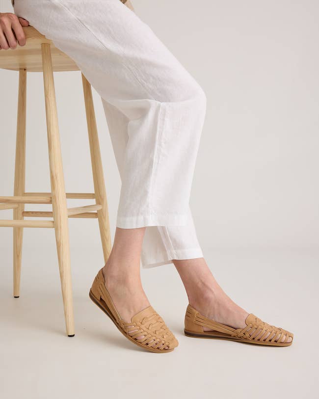 Person wearing light loafers and white pants sits on a wooden stool, showcasing footwear. Ideal for casual shopping insights