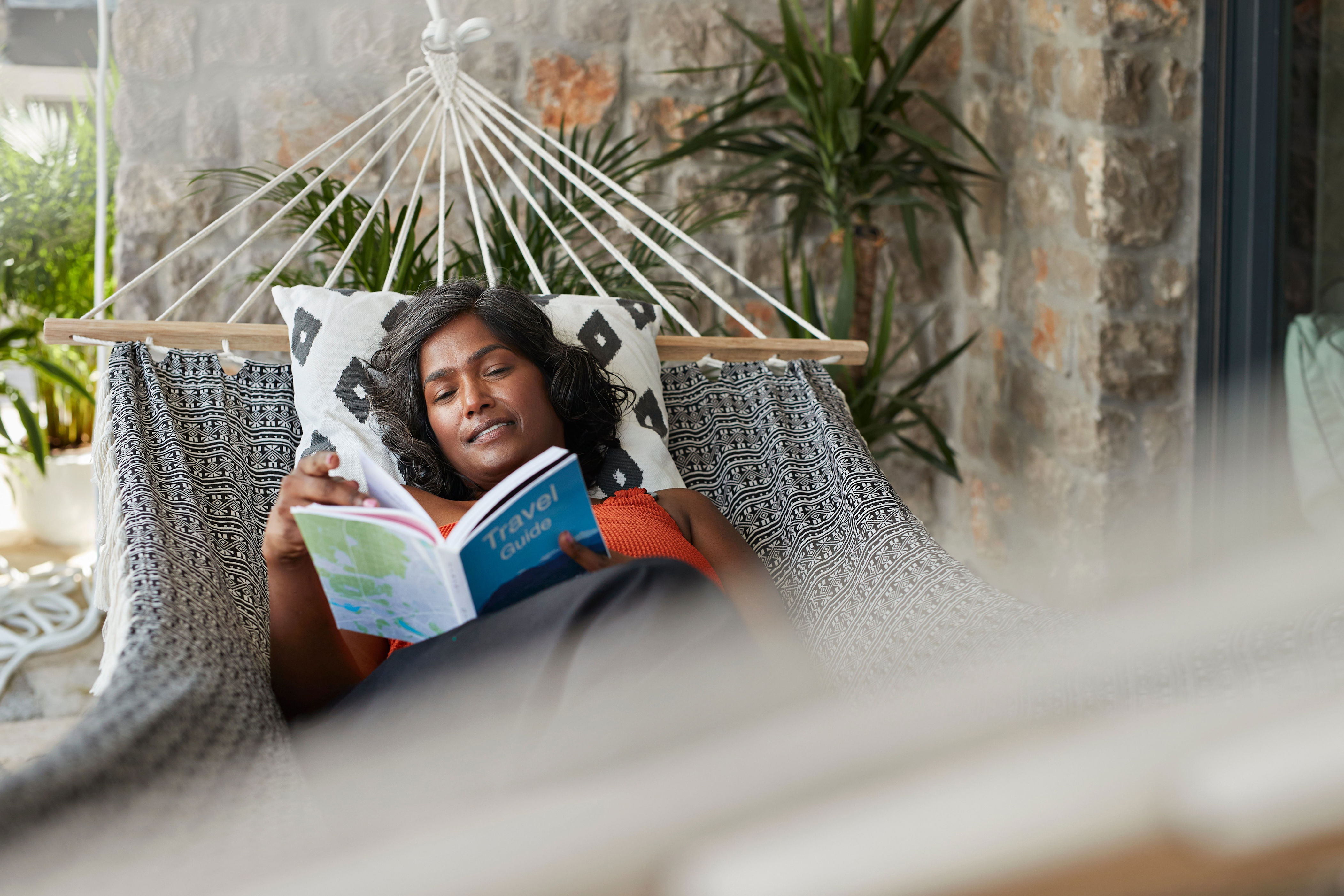 Person relaxing on a hammock, reading a travel guide. Brick wall and plants in the background, suggesting a calm outdoor setting