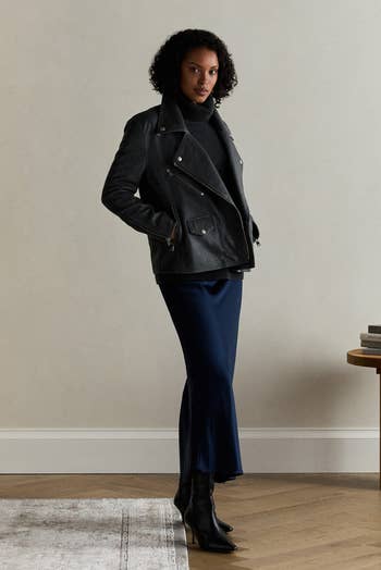 Person in a dark jacket and long skirt stands indoors on wooden flooring near a table with stacked books and fruit