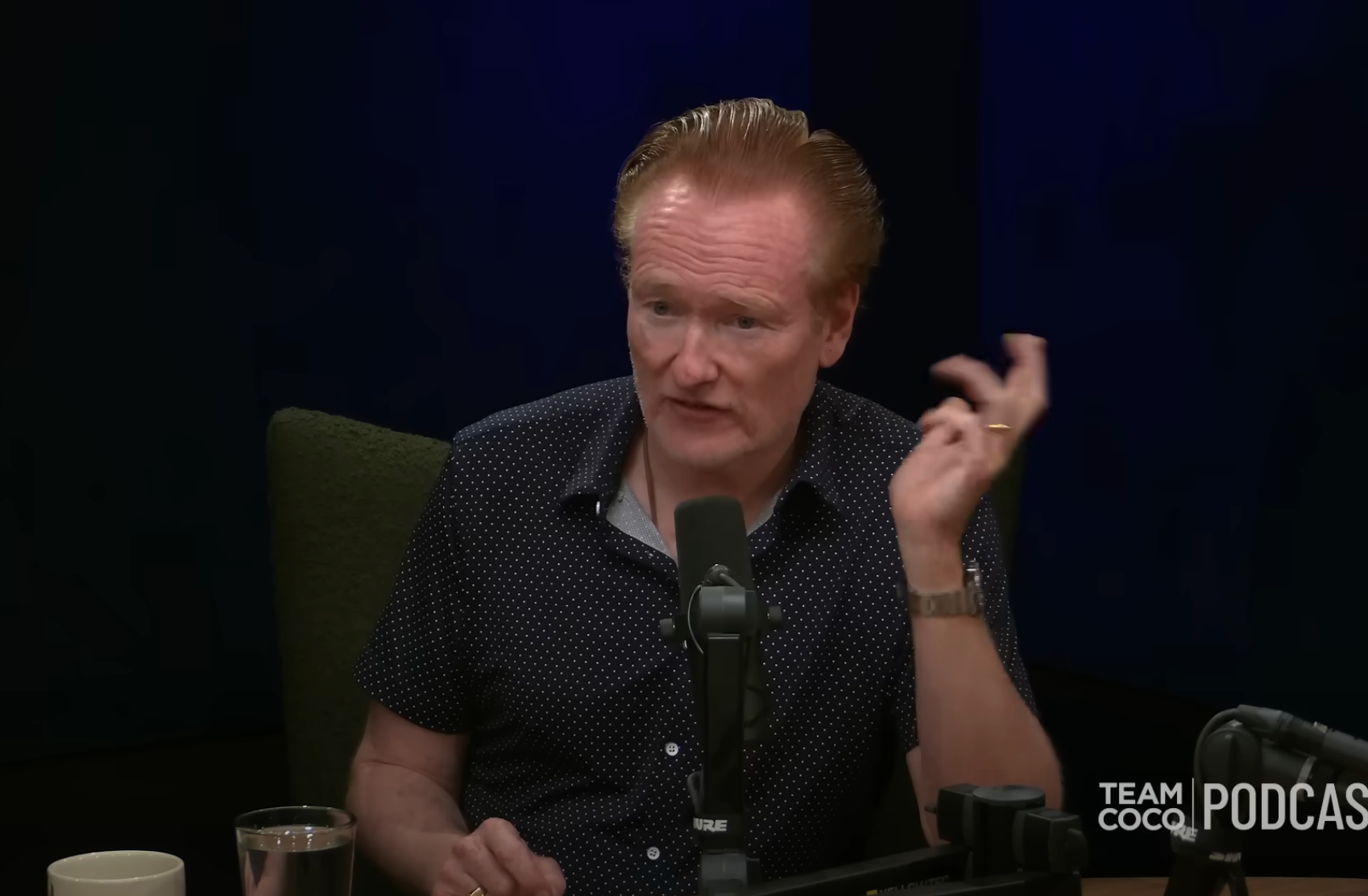 A person is speaking into a microphone at a podcast recording, with the words "Team Coco Podcast" displayed on screen