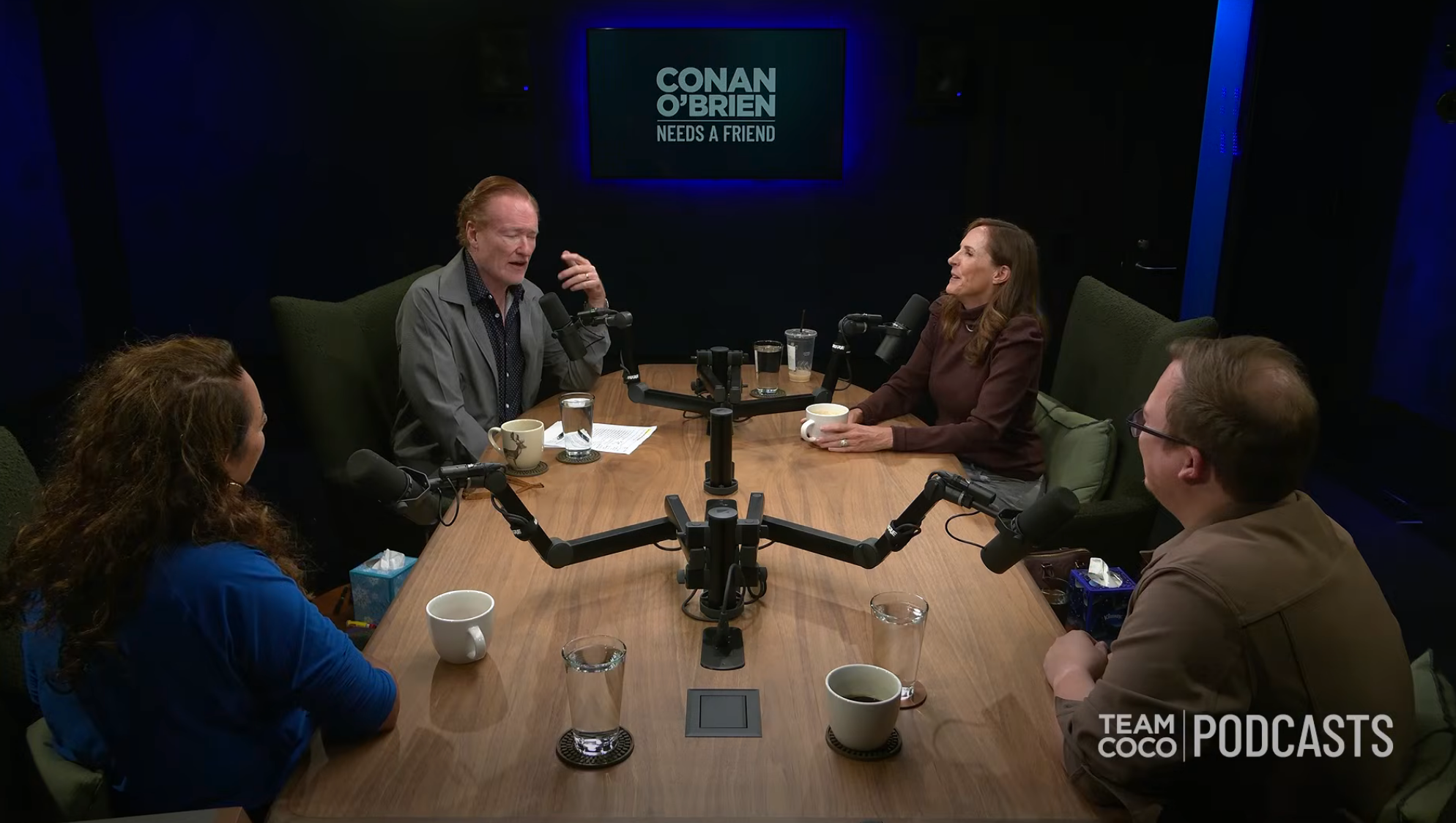 Four people sit around a podcast table with microphones, engaged in conversation on
