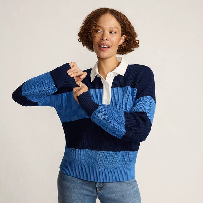Person wearing a striped sweater with a collared shirt underneath and jeans, posing with hands near face