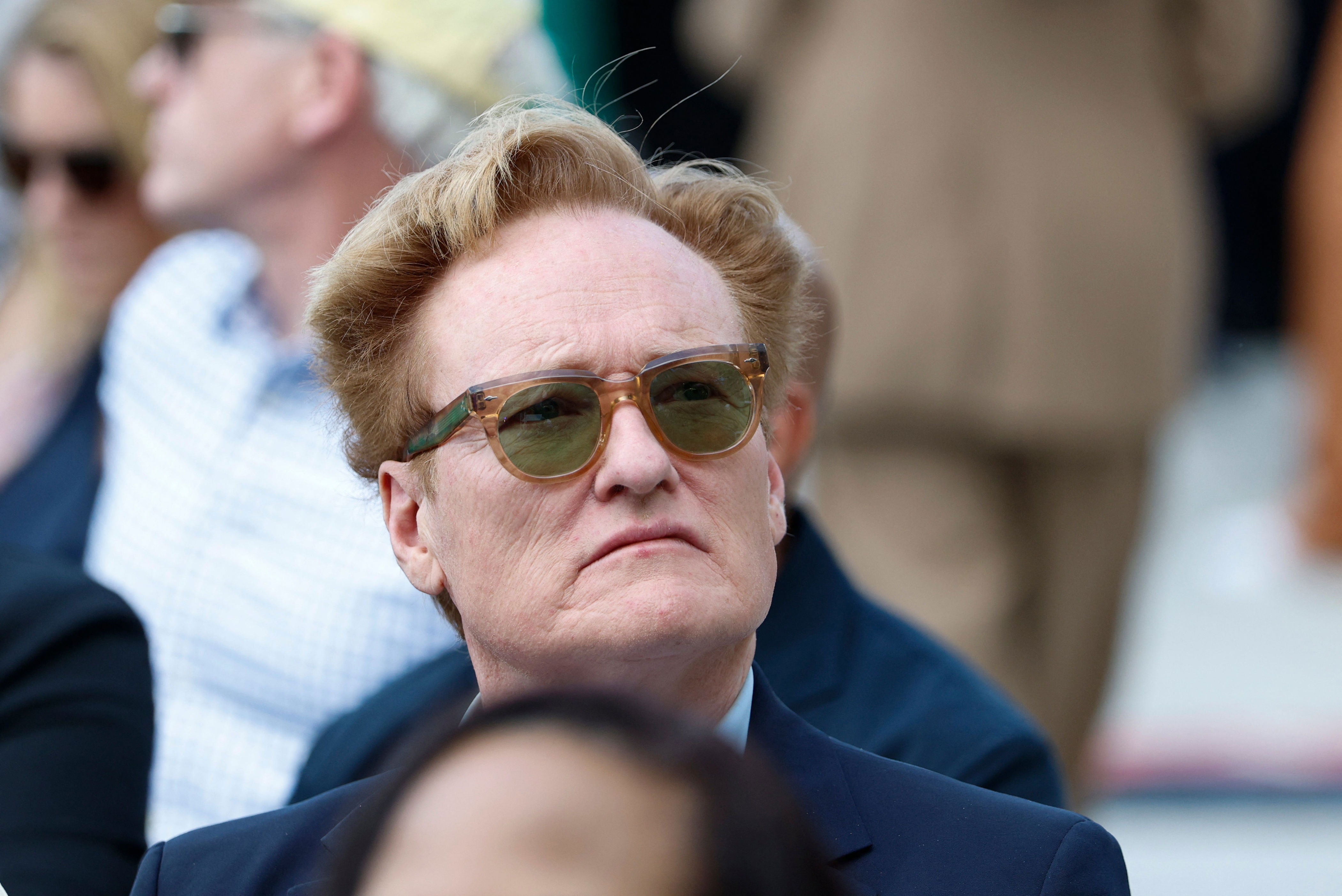 Person wearing sunglasses and a suit jacket, looking ahead in an outdoor setting with people in the background