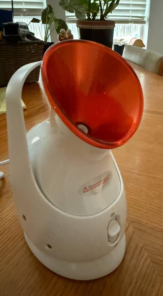 Facial steamer on a wooden table, featuring a white base and a wide red nozzle, used for skincare and wellness routines