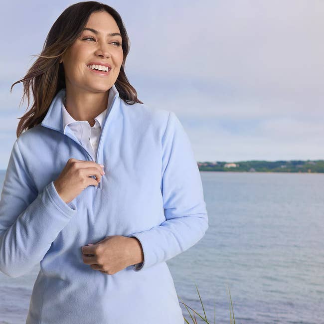 Person smiling near a lake, wearing a light jacket over a collared shirt, suitable for casual outdoor activities