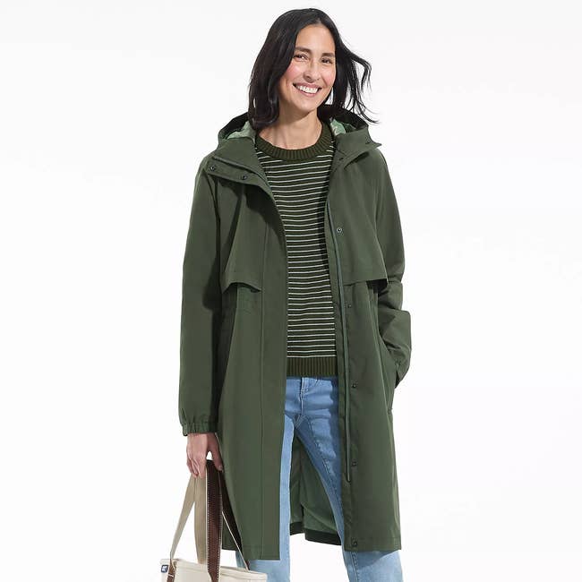 A person smiling, wearing a long, casual green coat over a striped sweater and jeans, holding a tote bag, suitable for a shopping context
