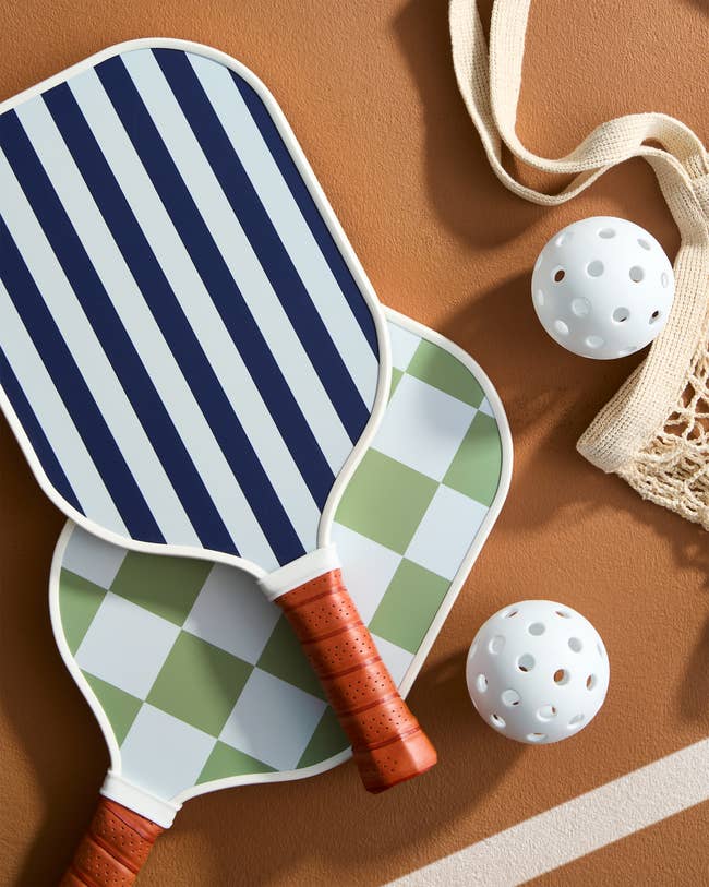 Two pickleball paddles with striped and checkered designs, two perforated balls, and a net bag on a court surface