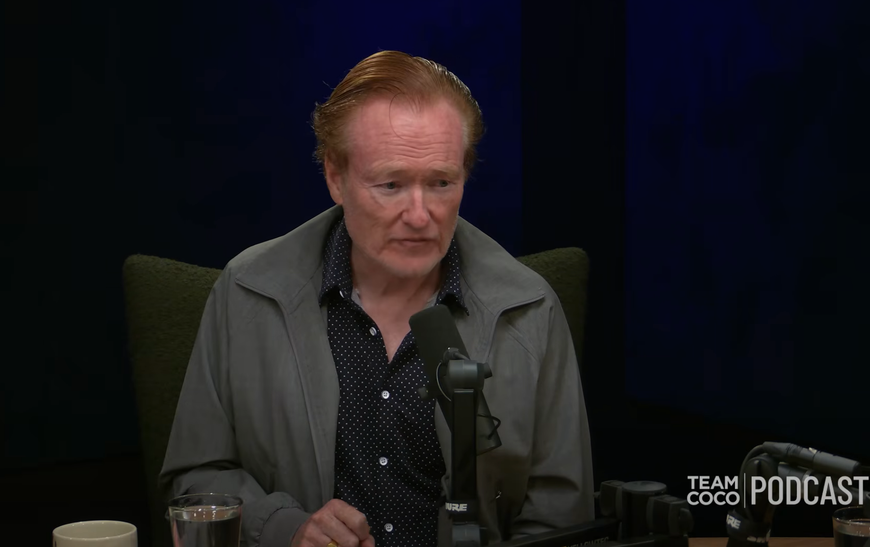 A person is sitting in front of a microphone on a podcast set, wearing a polka-dotted shirt and a jacket