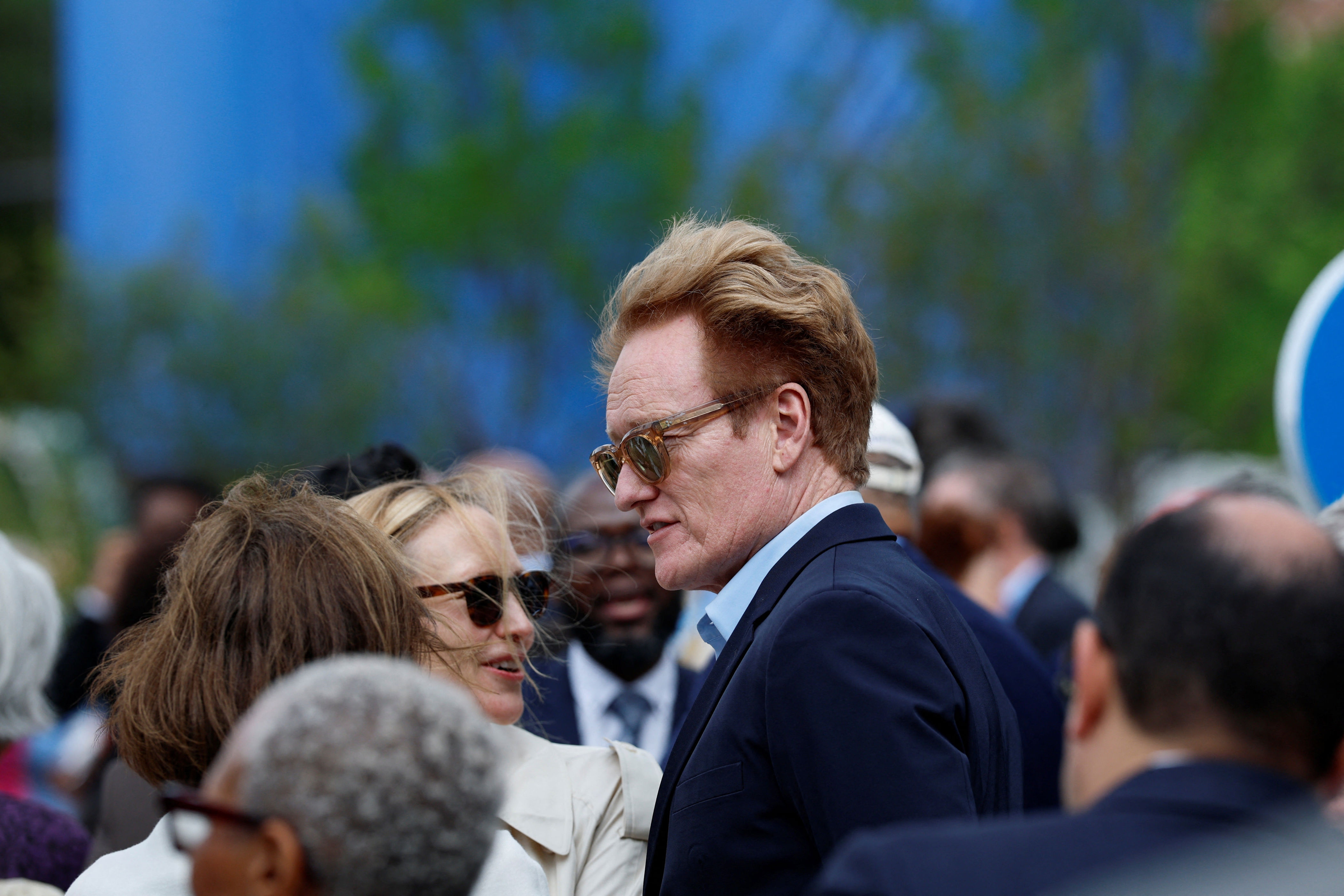 A man with styled hair and sunglasses wearing a suit is talking to a woman in a crowded outdoor setting