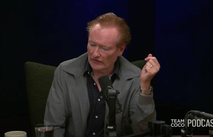 Person speaking into a microphone during a podcast, wearing a casual button-up shirt with a jacket