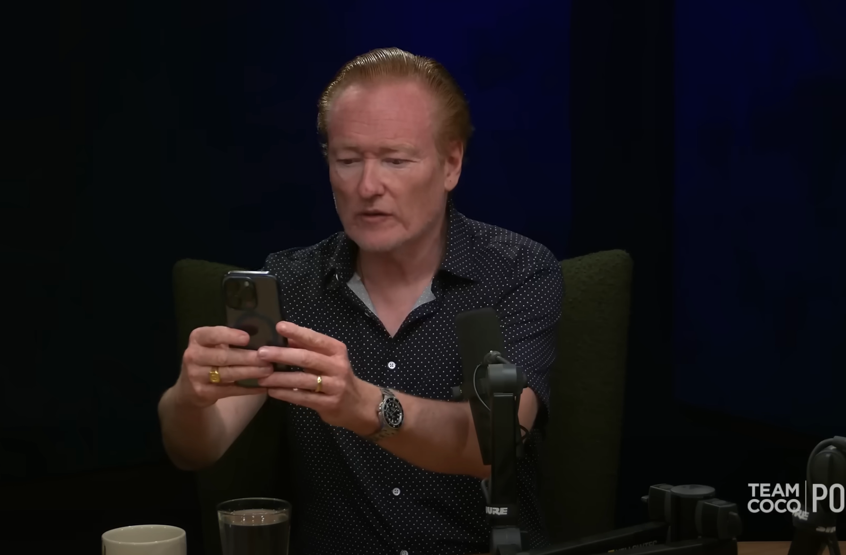 Person in a podcast setting, holding a smartphone, appears engaged. Microphones and drinks are visible on the table