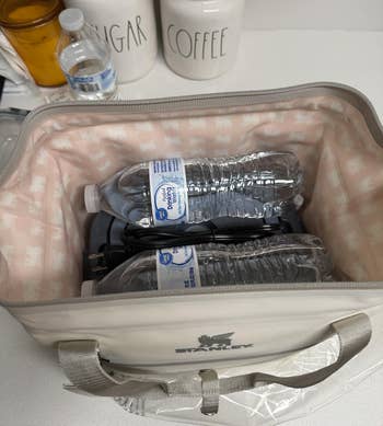 Open cooler bag with water bottles and a phone charger inside, placed on a kitchen counter next to sugar and coffee containers