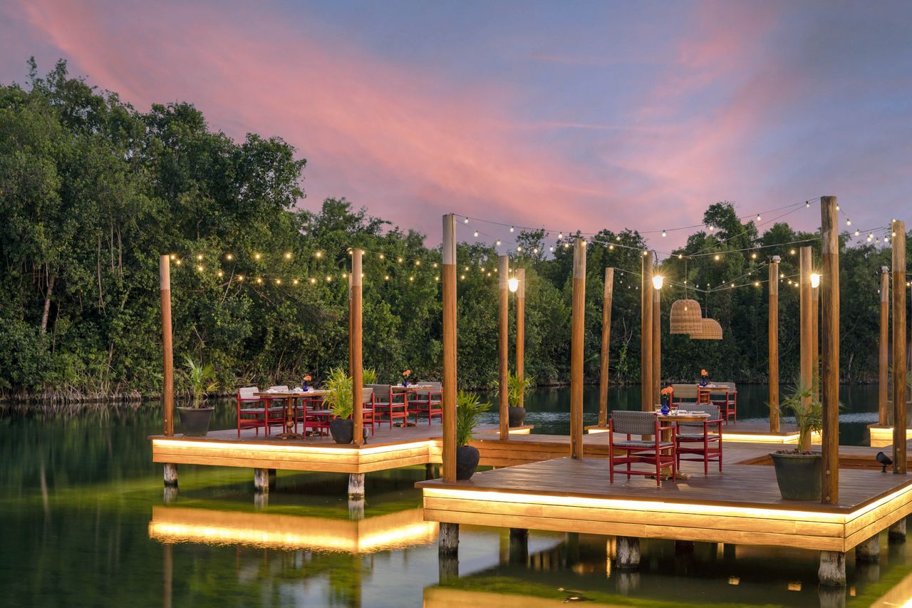 Outdoor wooden dining decks over water with string lights surrounded by greenery during sunset
