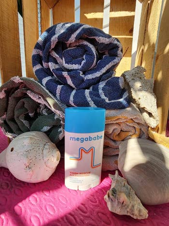 Megababe Thigh Rescue stick displayed with rolled towels and seashells on a pink surface