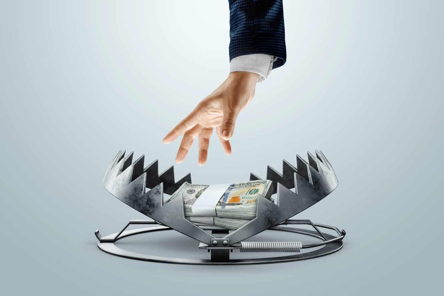 A human hand in a suit sleeve reaches downwards towards a stack of banded US hundred-dollar bills. The money is placed on the bait plate of a large, menacing metal bear trap that is set open, ready to spring. The background is a clean, solid light grey.