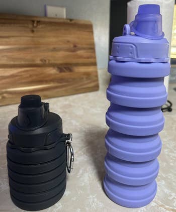 Two collapsible water bottles on a kitchen counter; one is purple and extended, the other is black and compact