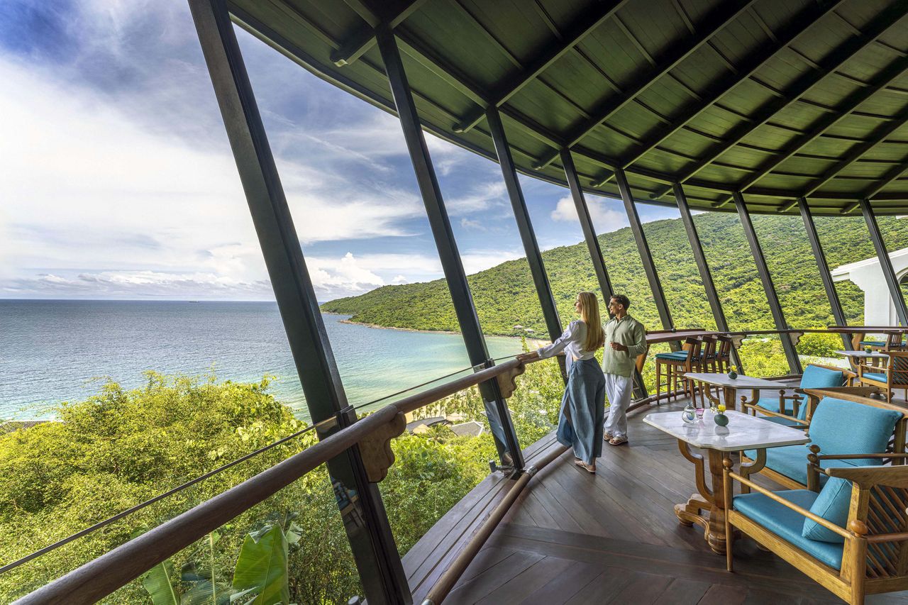 People on a terrace overlooking a seaside and forested landscape featuring lounge chairs and a table