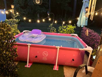 An inflatable pool with a purple float is on a patio with string lights, surrounded by plants and a garden umbrella