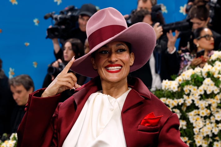 Person in a stylish outfit with a wide-brimmed hat and a red coat, smiling and posing on a floral-decorated event carpet
