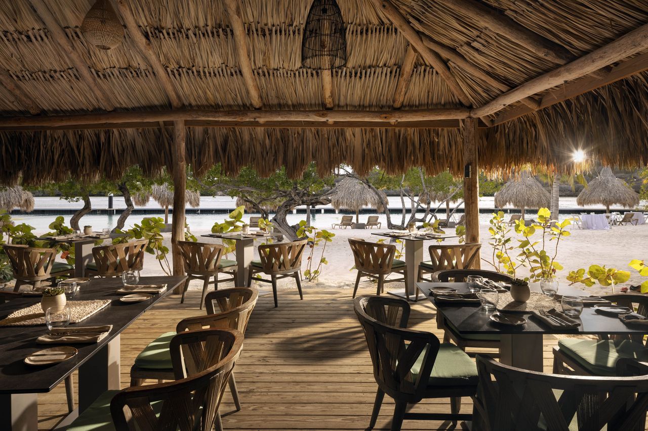 Outdoor restaurant area with tables and chairs under a thatched roof by the beach