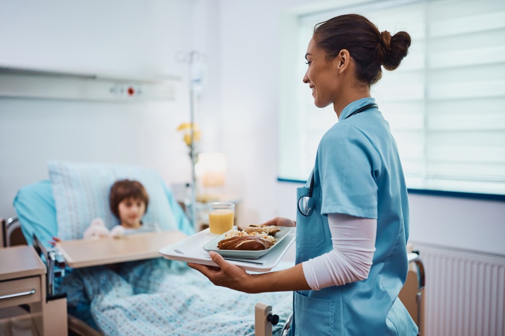 Young pediatric nurse serving lunch to a child at hospital ward.