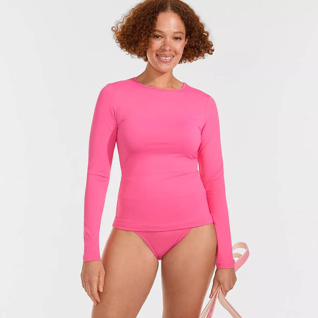 Person smiling in a pink long-sleeve swim top and bikini bottoms, holding a ribbon, suitable for a shopping article