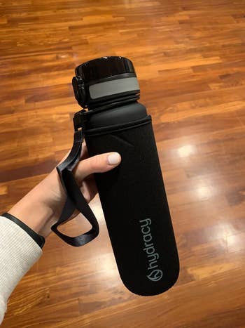 Person holding a black Hydracy water bottle with a strap, set against a wooden floor